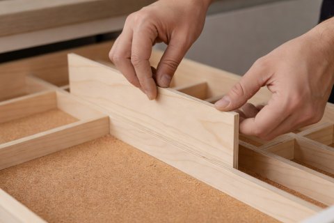 Hands fitting a sanded birch plywood divider strip into a cork-lined kitchen drawer